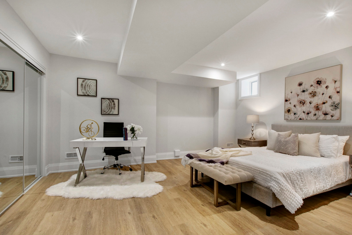 Large master bedroom with white ceiling and wooden floors staged for sale with king bed, desk, office chair, white fur carpet, wall painting, side table and lamp.