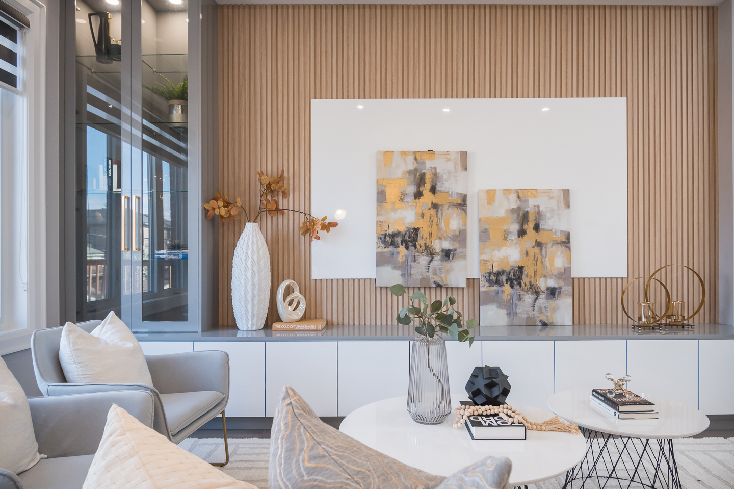 Bright contemporary living room featuring a beige sofa, neutral decor, wood slat accent wall with abstract artwork, built-in cabinetry, and large window with natural light.