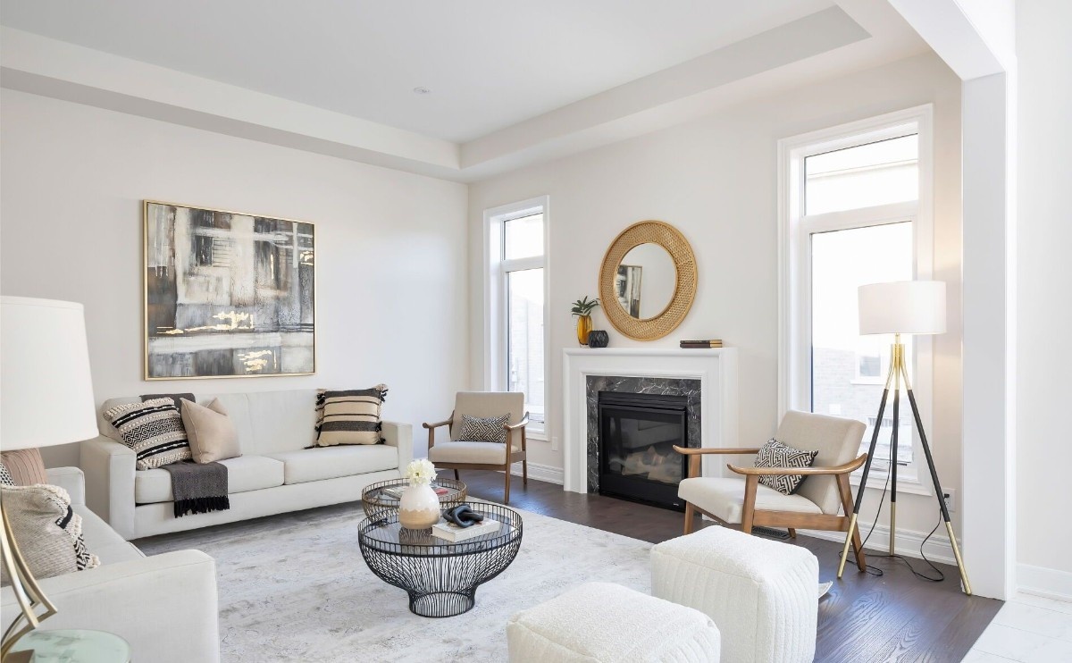 Bright, fully staged luxury living room featuring a white sofa, accent chairs, round coffee tables, wall art, a fireplace with mirror above, soft neutral tones, and modern lighting.