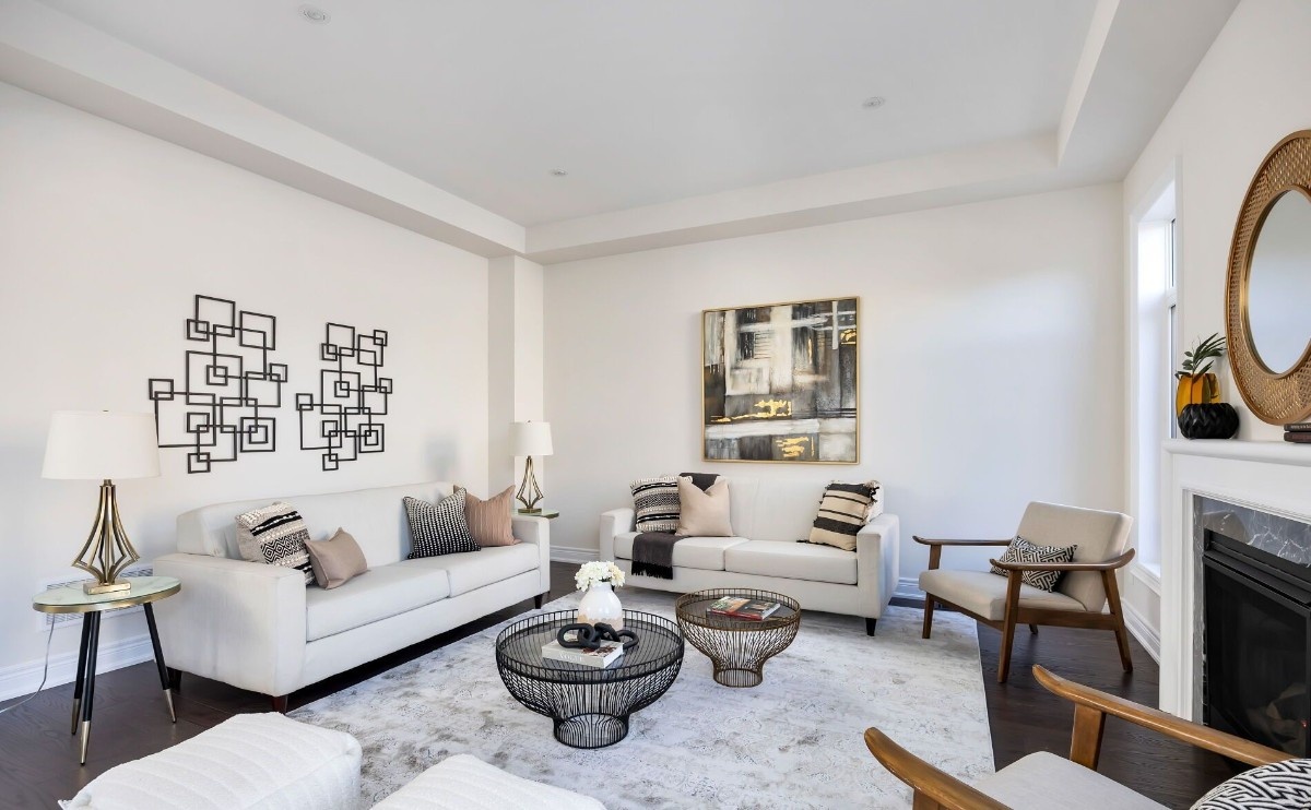 A stylish living room featuring two cream-colored sofas arranged around round wire-frame coffee tables on a light area rug. The space includes modern wall art with geometric metal designs, a large abstract painting, and coordinated throw pillows. A side table with a lamp, a wooden accent chair, and a fireplace with decorative pieces complete the bright, neutral-toned interior.