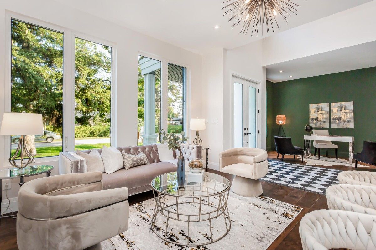 A sunlit living room featuring plush beige seating, including a sofa and curved armchairs arranged around a glass coffee table on a patterned rug. Large floor-to-ceiling windows bring in natural light and views of greenery outside. In the background, a stylish workspace with a dark green accent wall, desk, and modern lighting adds contrast to the bright, neutral-toned interior.