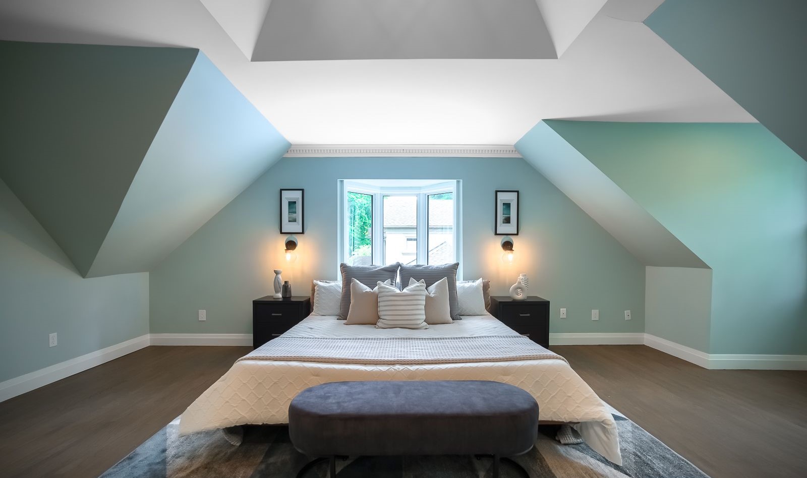 Staged attic bedroom with sloped ceilings, teal accent walls, centered bed, nightstands with lamps, and a window providing natural light.